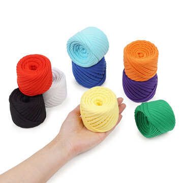 Close-up product photography of a hand gently holding a yellow cotton yarn skein. Multiple colorful thick yarn rolls on clean white background.