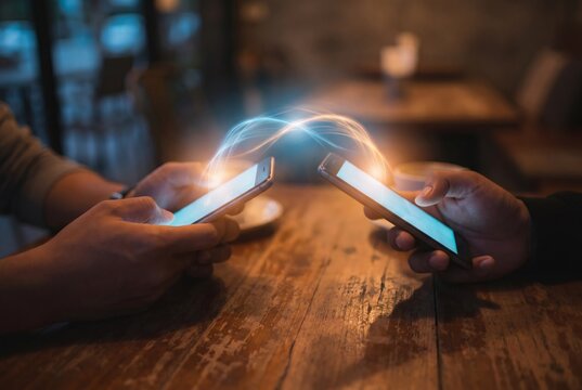 Two hands holding smartphones with glowing light connection transferring data on wooden table in cafe, peer to peer payment and mobile technology concept