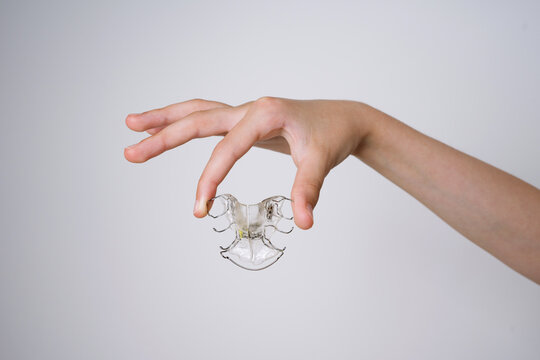 Child holding a removable orthodontic retainer plate on a white background. A close up of a child's hand holding a removable clear plastic orthodontic retainer plate with metal wires