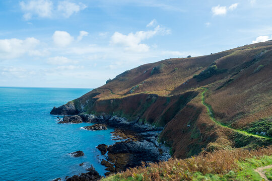 Dramatic coastal path winding along rugged cliffs above turquoise sea, grassy slopes and rocky shoreline under a bright sky. Jersey, Channel Islands, UK.