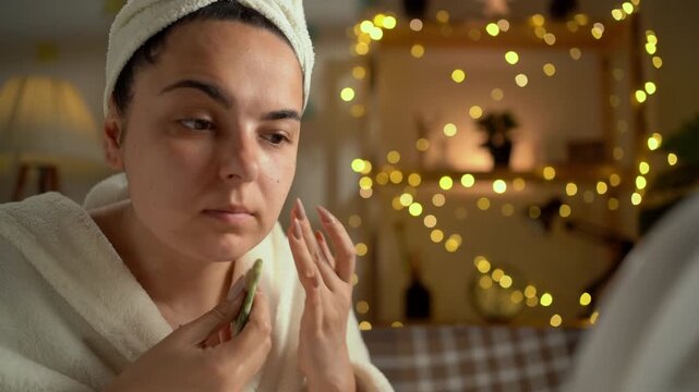 close-up portrait Caucasian woman in bathrobe enjoying her skincare routine, looking in mirror, using a jade gua sha stone to smooth out the skin on her face, creating a relaxing atmosphere at home