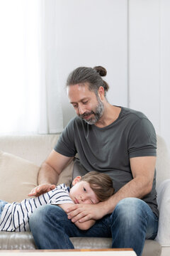 A father sits on a couch holding his son who is sleeping. The child is lying down with his head on his father's lap