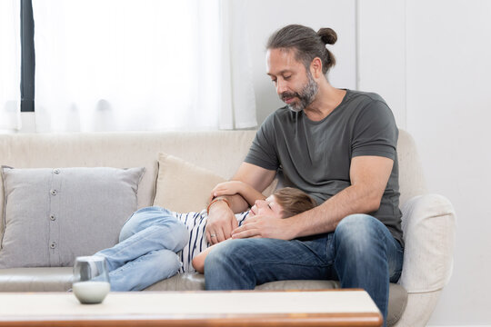 A father sits on a couch holding his son who is sleeping. The child is lying down with his head on his father's lap