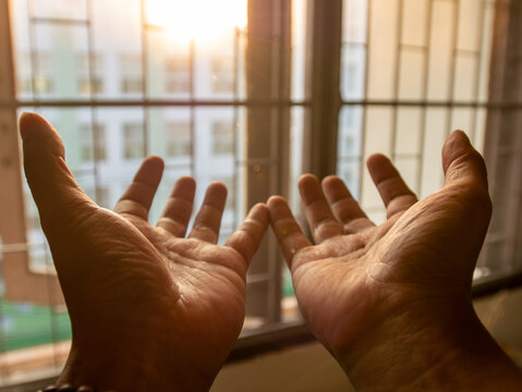 Two open hands palms facing up raised toward sunlight through window with warm golden backlight spiritual prayer concept