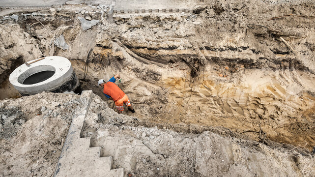 A worker is digging in a deep pit to construct a concrete sewer well. The area is filled with dirt and debris, showing ongoing construction work