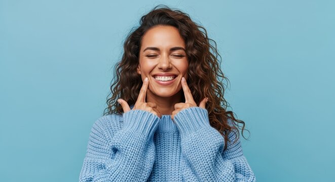 Happy young woman with curly hair pointing at her beautiful smile and dimples on blue background