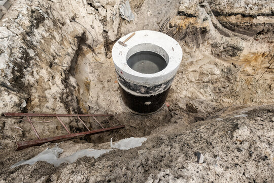 Workers are building a concrete sewer well in a deep pit at a construction site. Tools and a ladder are present near the well during daytime