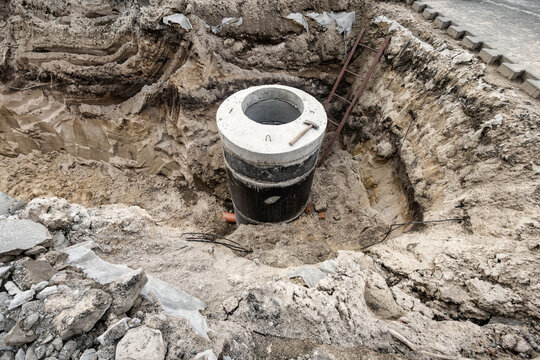 Workers are constructing a concrete sewer well in a deep pit at a construction site. Tools and materials are present in the area
