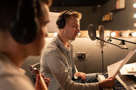 Male Singer reading song lyrics near guitarist in recording studio 