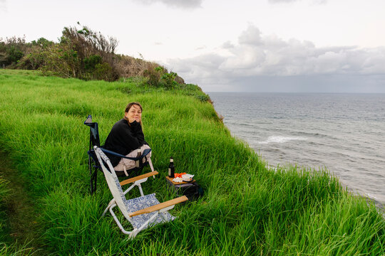 UGC woman sitting by coastal picnic setup on grassy cliff at sunset