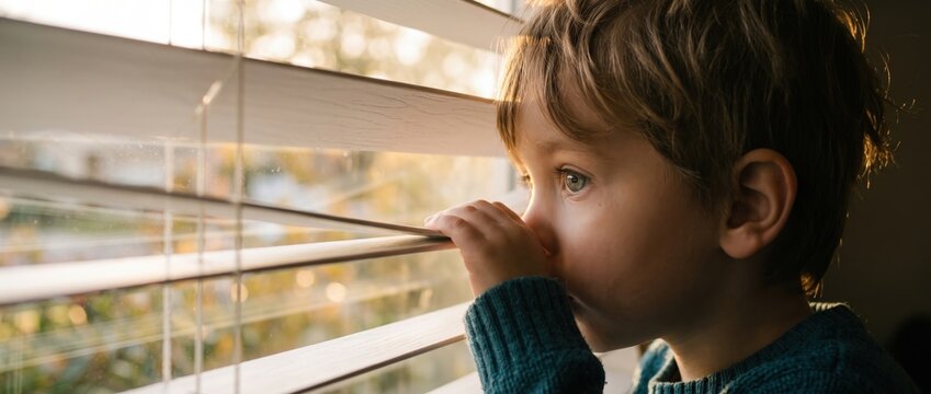 Young boy looking through window blinds at golden hour light with curious expression and soft focus background in a warm home interior setting