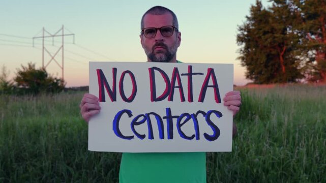 Middle-aged man holding handmade No Data Centers placard peaceful protest in Midwestern United Stated rural field near industrial area