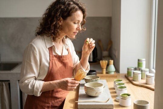 Woman in an apron holding a matcha whisk, preparing green tea ritual in a kitchen