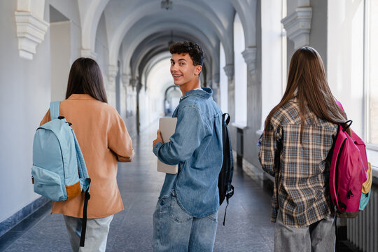 Back view of high school students walking down school corridor, teenage classmates going to class, one boy turning back and looking at camera, education and student life concept