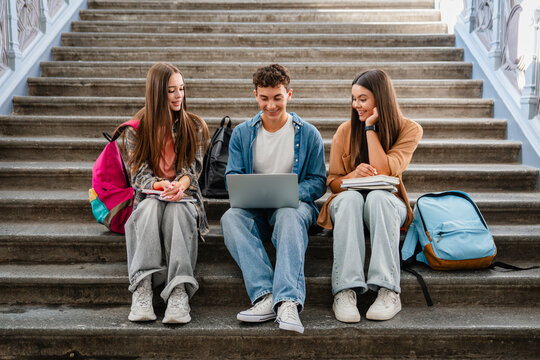 Group of high school students sitting on campus stairs with textbooks and laptop, classmates talking together, digital learning, education and student life concept