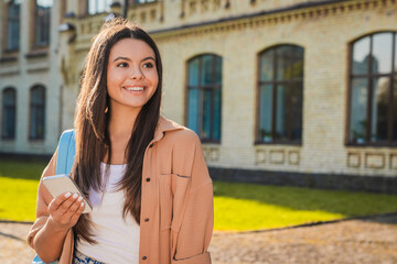 Fototapeta premium Close up of female student holding smartphone and looking at copy space in front of university building, mobile internet, online learning, digital communication and student life concept