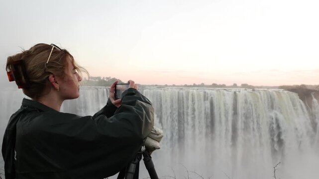 young woman taking pictures of sunrise over Victoria Falls from the Zimbabwe side, Zimbabwe, Africa