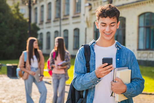 Young male student holding smartphone near campus building with group of students in background, mobile internet, education and modern student lifestyle concept