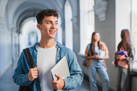 Portrait of male student with laptop in school corridor, classmates talking in background, modern education, digital learning and student life concept