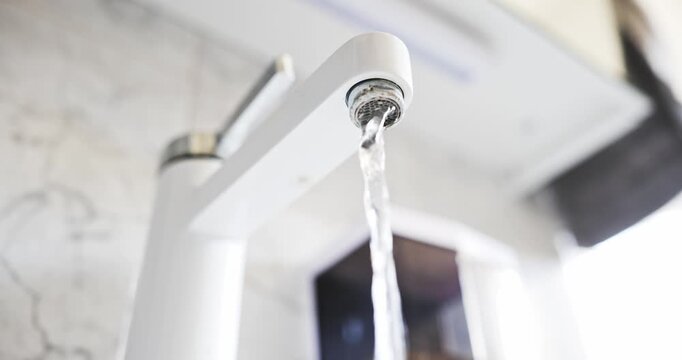 Water flowing continuously from the calcified aerator of a modern white bathroom faucet in a low angle shot in real time.