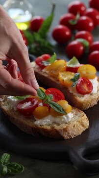 Close-up vertical video of a hand putting basil leaves on toasted Italian bread, or bruschetta, with creamy stracciatella cheese and red and yellow cherry tomatoes. Mediterranean appetizer, homemade I