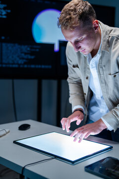 A focused blond cybersecurity analyst interacts with a glowing touchscreen panel at a glowing command-centre workstation at night, tracking suspicious activity across enterprise systems.