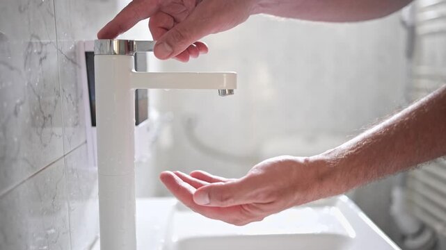 Man turning on a modern white faucet, cupping water in his hand, and letting it splash over in slow motion.