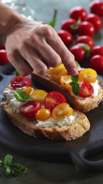 Close-up vertical video of a hand putting basil leaves on toasted Italian bread, or bruschetta, with creamy stracciatella cheese and red and yellow cherry tomatoes. Rustic Mediterranean appetizer, hom