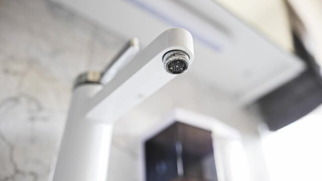 Water dripping slowly from the calcified aerator of a modern white bathroom faucet in a low angle shot in slow motion.