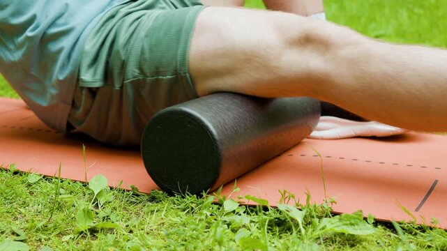 Close-up of a man doing foam rolling on his hamstring for post-workout recovery and self-massage. Myofascial release of muscle tension on a fitness mat in a park. High quality 4k footage