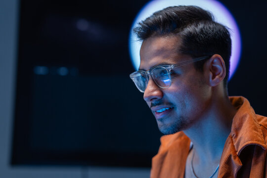 A focused dark-haired cybersecurity analyst smiles confidently at his workstation inside a dark security operations centre at night, tracking suspicious activity across enterprise systems.