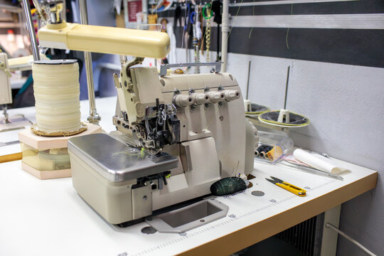 An industrial overlock sewing machine sits on a wooden workbench, surrounded by thread spools, scissors and tailoring tools in a workshop