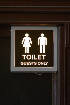 A square illuminated restroom sign features male and female pictograms above the text toilet guests only against a dark background.