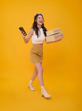 Asian woman walking with delivery boxes and smartphone representing mobile ecommerce order fulfillment customer service fast shipping digital retail and joyful online shopping.