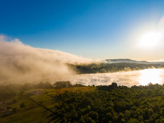 Fototapeta premium Morning fog covers a river while the sun rises over the hills and illuminates the landscape in a rural area near a bridge