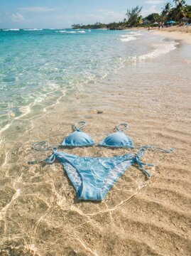 A beautiful vertical lifestyle shot of a light blue textured two-piece string bikini submerged in shallow, crystal-clear ocean water on a pristine sandy beach.