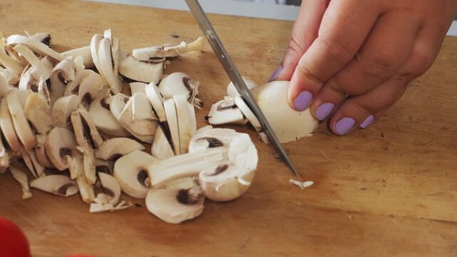 A woman's hands slice champignons on a cutting board and toss them into a plate. Medium shot.