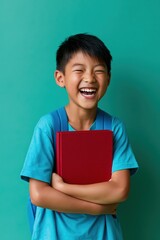 Obraz premium Laughing schoolboy holding red book against green background for education poster and children learning concept