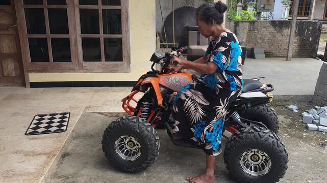 An adult woman in a floral dress getting onto an orange quad bike parked outside a house