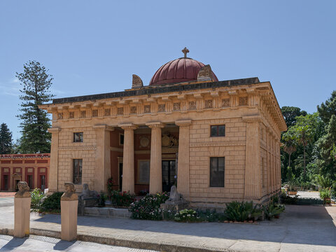 Neoclassical Gymnasium building at the Botanical Garden of Palermo, Sicily. Historic Ginnasio pavilion with a red dome and columns surrounded by lush palm trees under a blue sky.