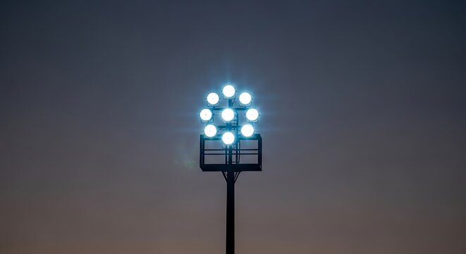 A lone stadium floodlight tower shines bright against a fading day sky