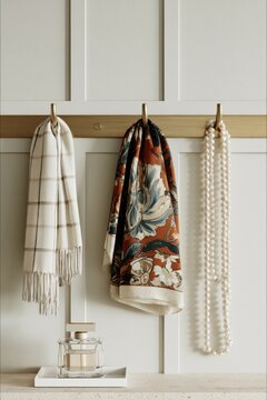 Closeup of a brushed brass hook rail displaying a patterned scarf, a checkered throw, and a string of pearls against a cream-painted wall with paneling