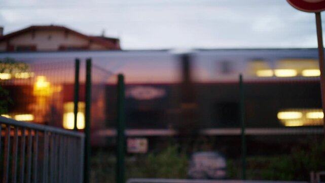 Soft focus passenger train passing behind a fence at dusk near Antibes, France, with glowing interior lights and a traffic restriction sign in the foreground