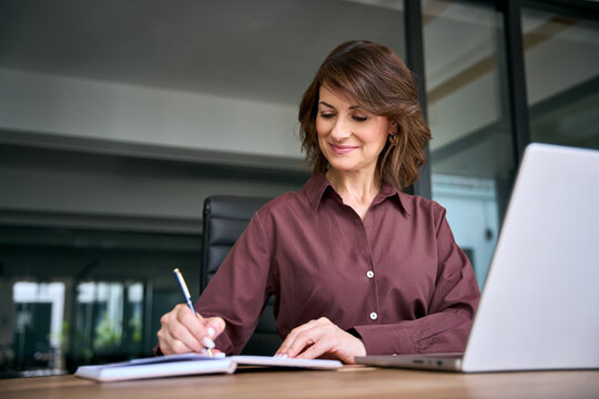Hispanic mature businesswoman writing notes in notebook at office desk. Woman executive holds pen, makes to do list, planning business schedule near laptop. Female manager organizes working day.