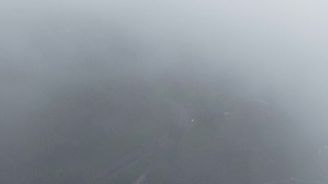 Aerial view of a winding mountain road barely visible through thick morning fog and mist over a dense forest, evoking solitude, mystery, calm and atmospheric landscape at dawn