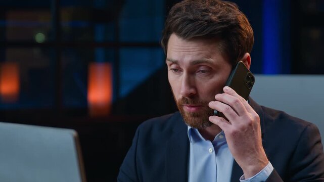 Focused middle-aged Caucasian business man talking phone in evening office worker looking in computer monitor working with desktop talk mobile call male businessman chatting with telephone at night