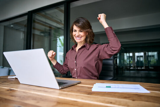 Excited hispanic businesswoman celebrating win at laptop in office. Happy mature woman trader raises fists, cheers victory and achievement. Overjoyed female winner gets good news, deal or profit.