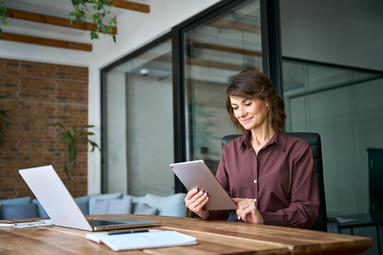 Working on digital tablet in modern office, latin mature businesswoman reads report online. Focused woman startup founder using device at desk. Female owner grows business company at workplace.