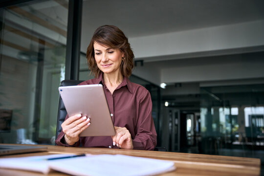 Businesswoman in 50s using digital tablet at office desk with copy space. Mature woman economist browses web on device, checks market email. Female business analyst reads reviews at workplace.