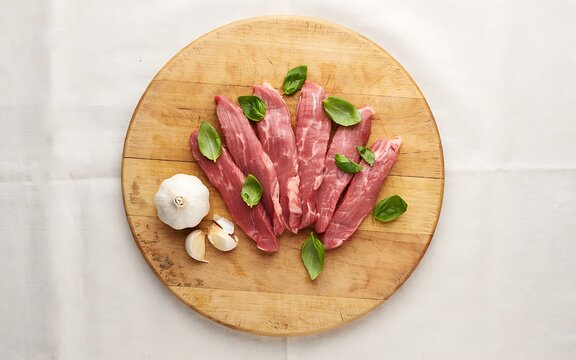 Freshly sliced raw beef steaks arranged on a wooden cutting board with garlic cloves and basil leaves for cooking preparation